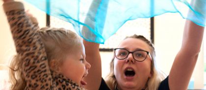 Learning through play at Kindred Huntingdon A teacher and a child raise a sheer blue scarf over their heads, their mouths open in a joyful expression.